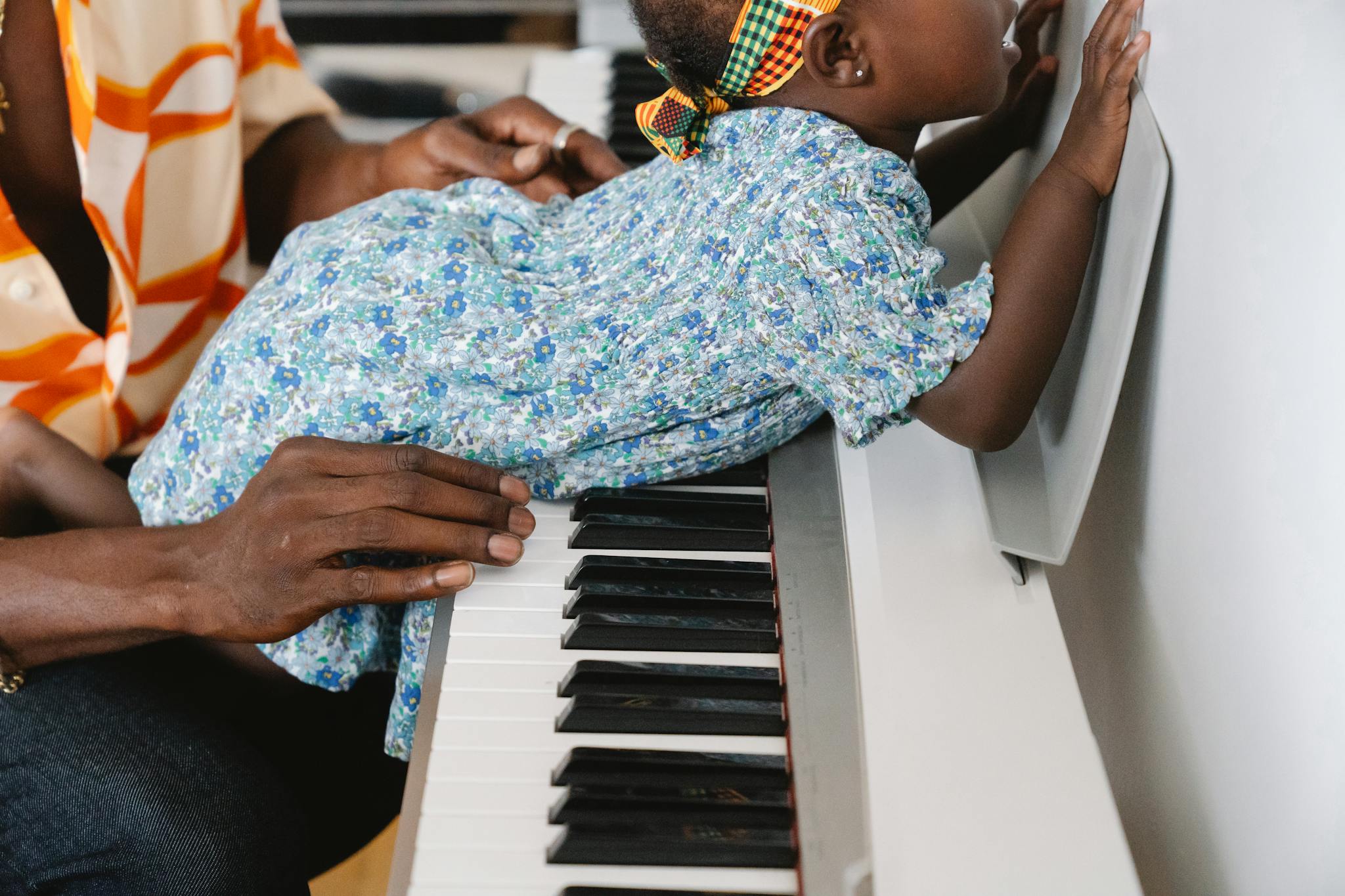 A baby in a floral blue dress interacts with a piano, assisted by an adult, showing curiosity and music exploration.